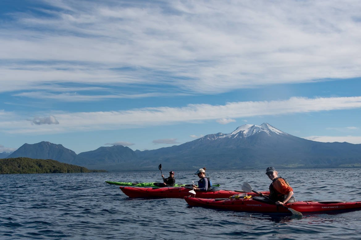Group in red and green sea kayaks on a wide lake with a snow-capped volcano behind