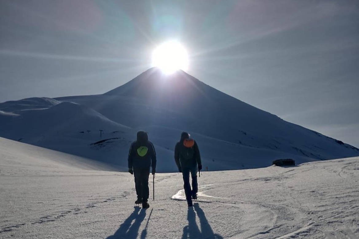 Dos personas caminan por la nieve hacia el cono de un volcán, con el sol detrás