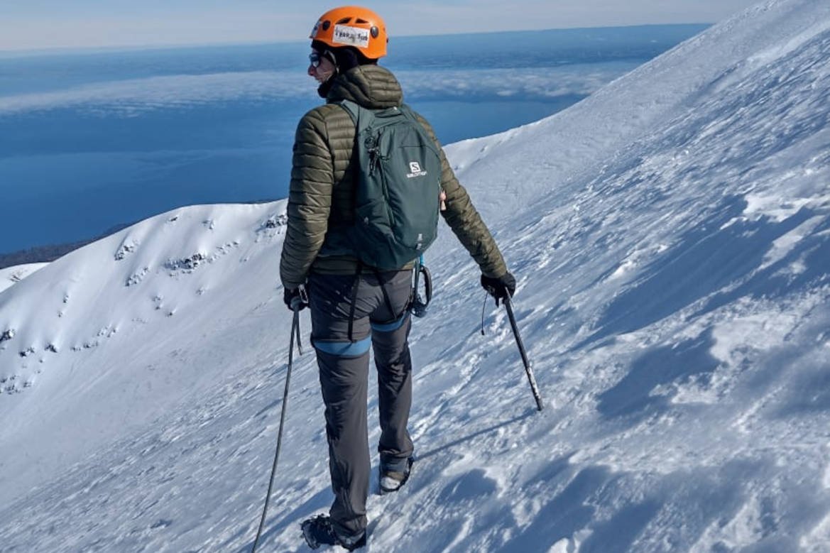 Persona con casco y bastones en una pendiente nevada, con un lago al fondo