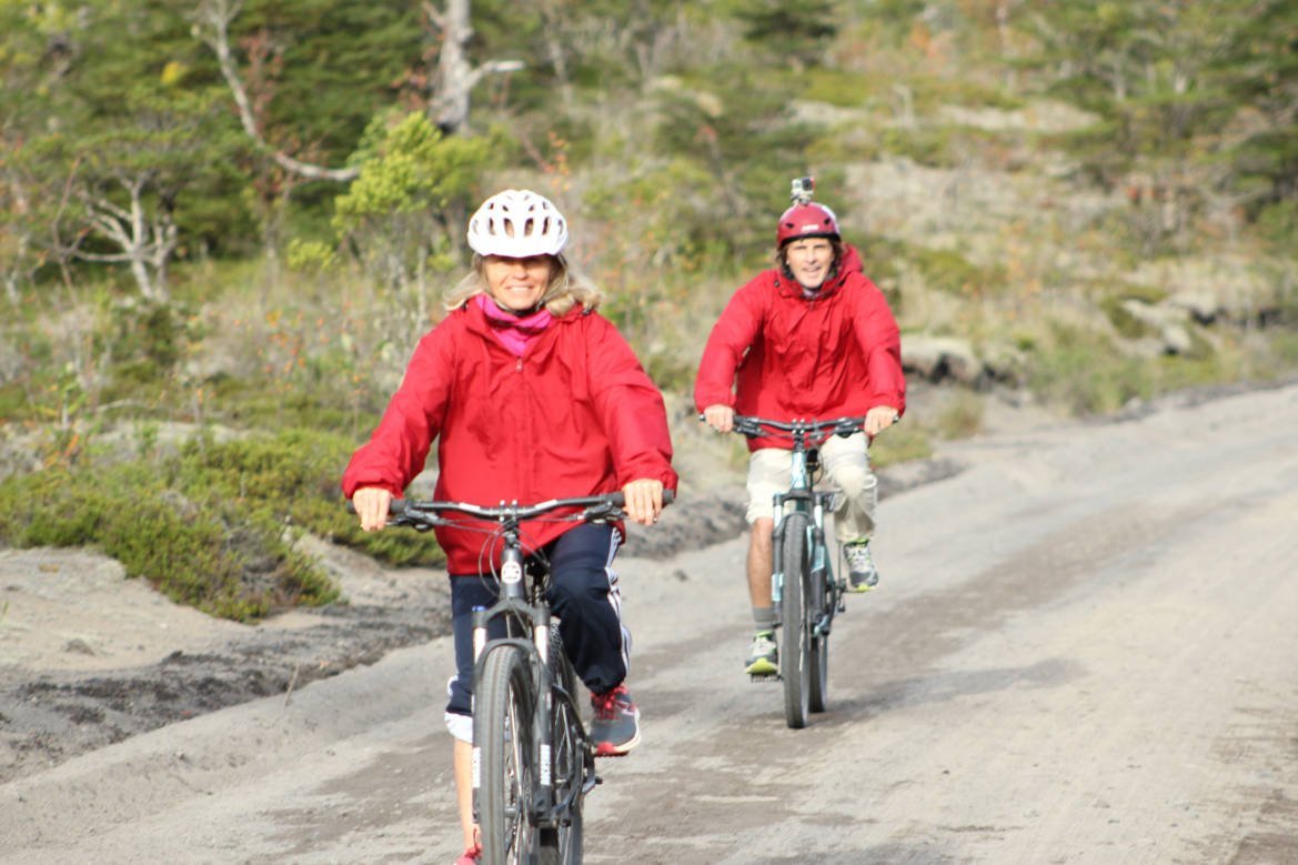 Two cyclists in helmets and red jackets riding down a gravel road among shrubs