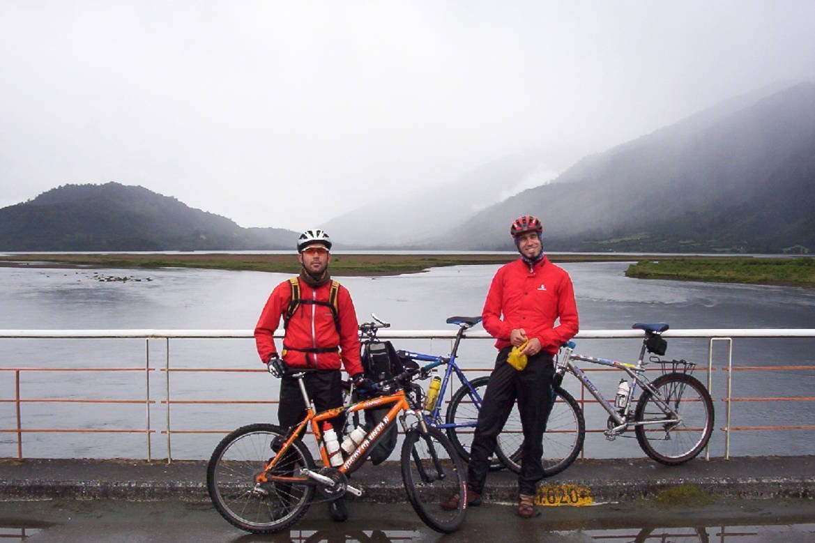 Dos ciclistas con sus bicicletas frente al agua y los cerros en un día nublado