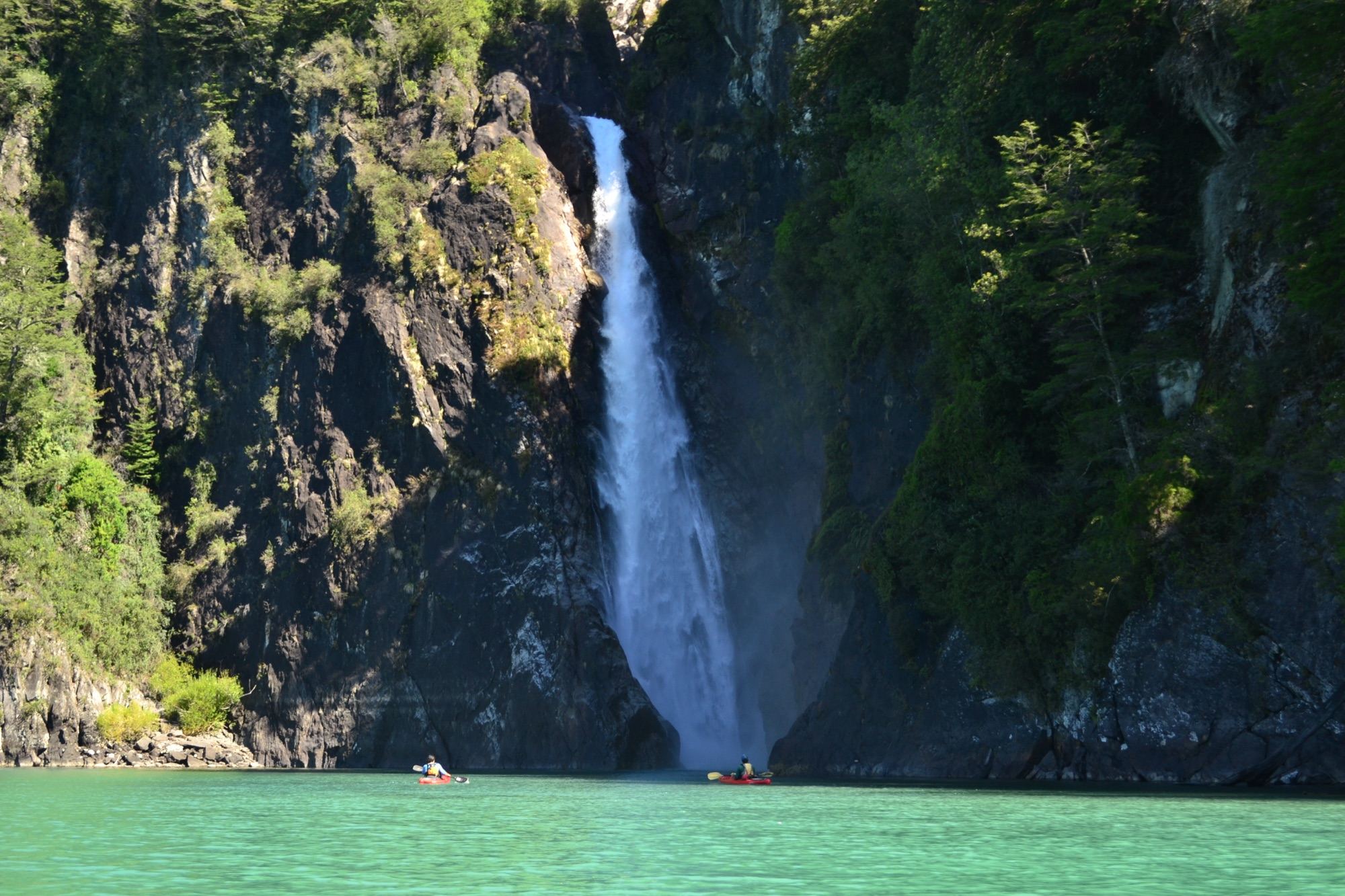 A waterfall drops between rock walls and forest into the turquoise water of Lake Tagua Tagua