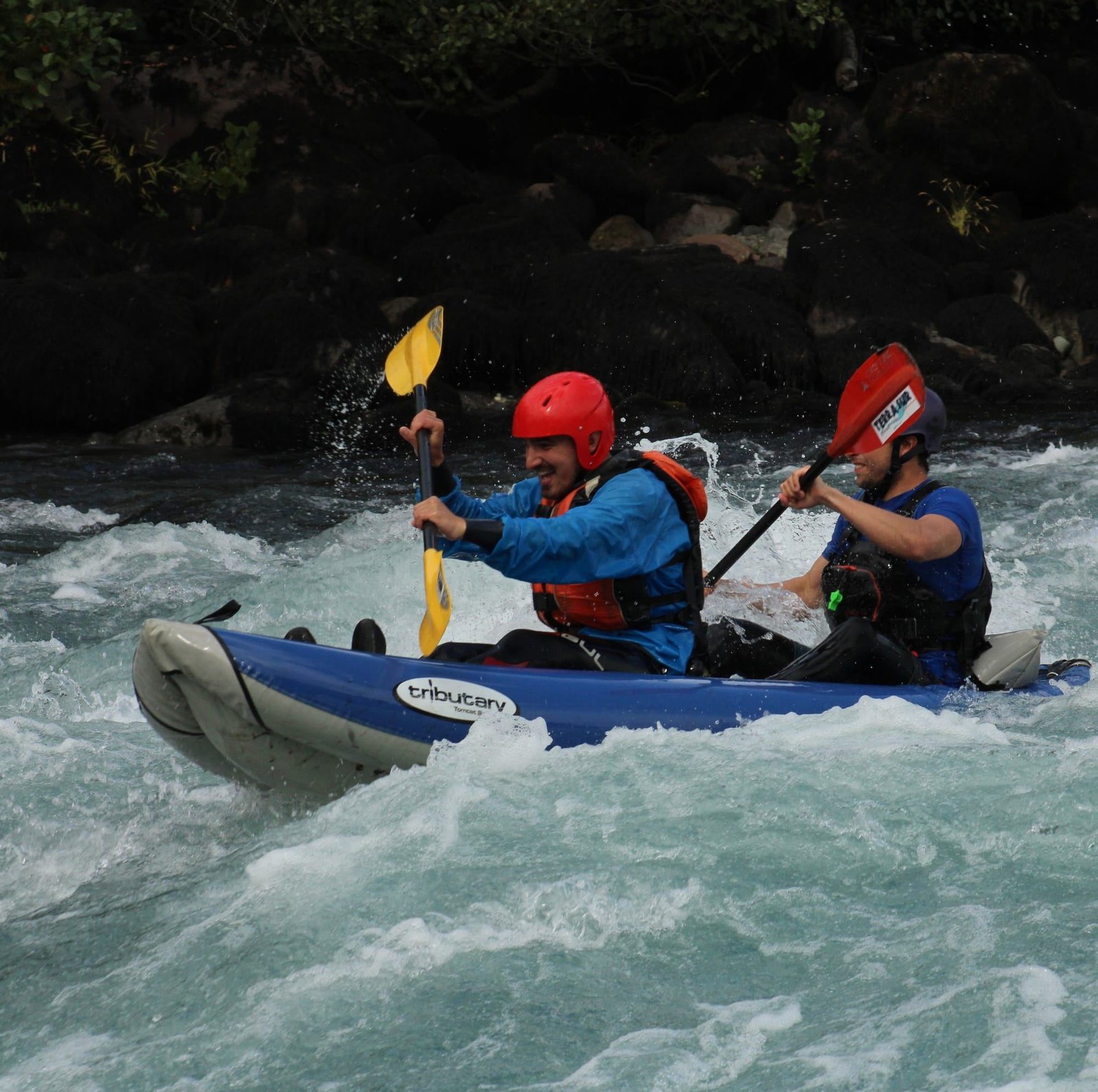 Dos personas con casco reman un kayak inflable azul entre olas