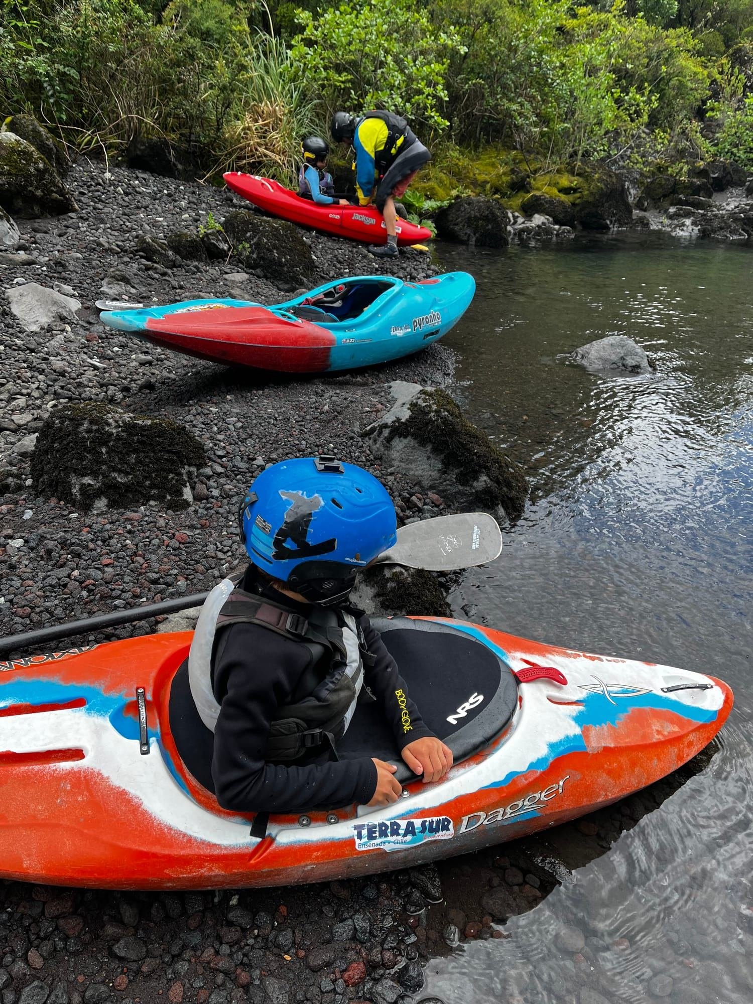 A person in a helmet sitting in a river kayak at the water's edge