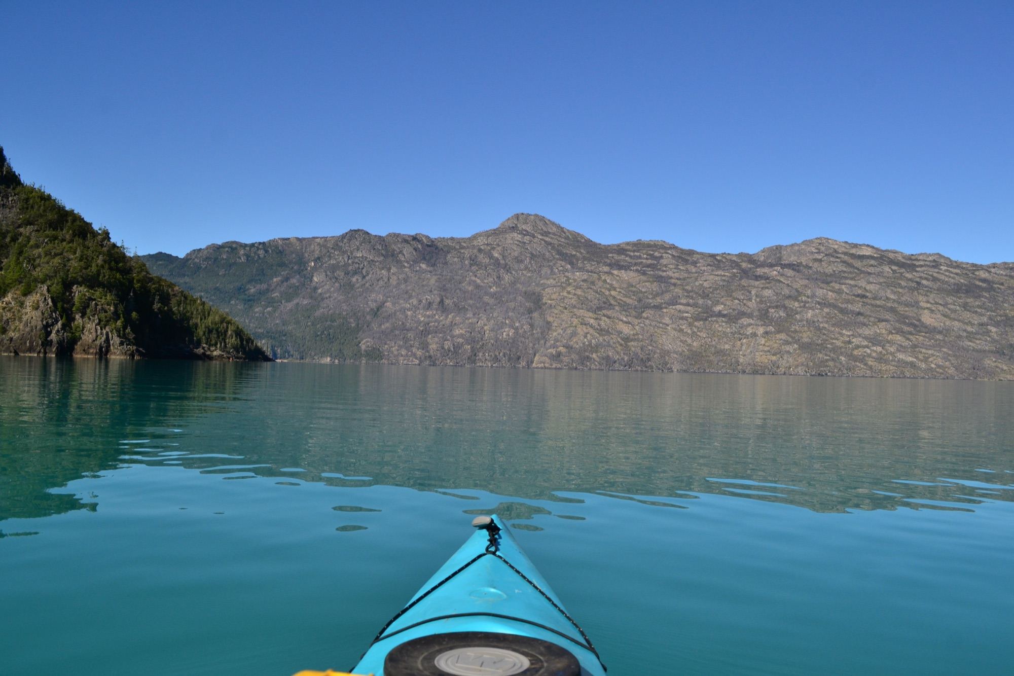 The bow of a light blue kayak on turquoise Lake Puelo, with hills behind