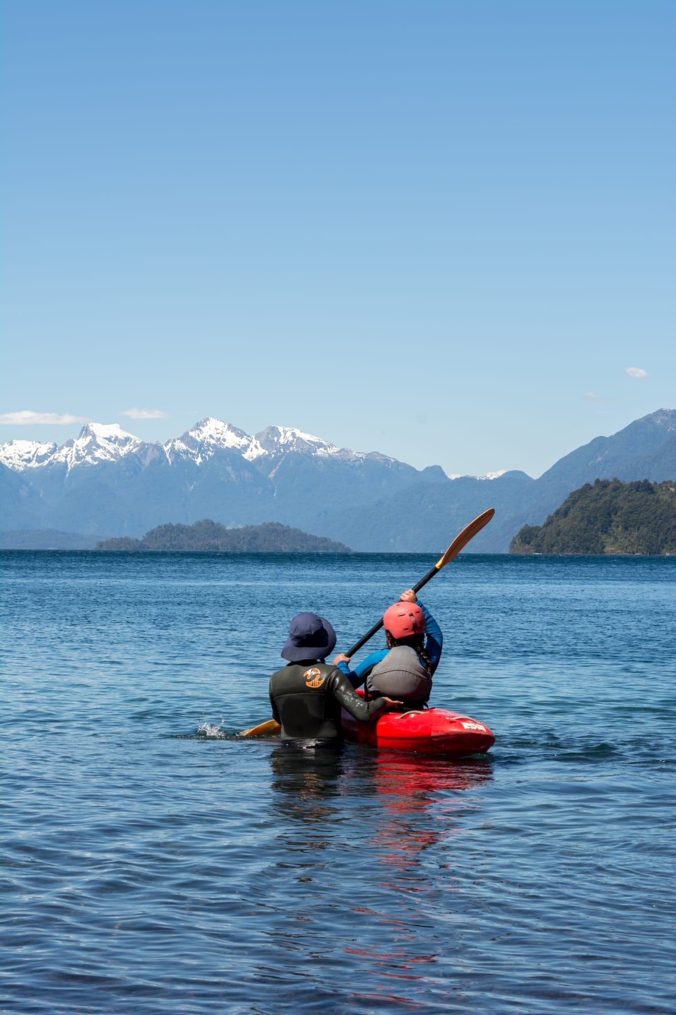 A person practices in a river kayak, with snowy mountains behind