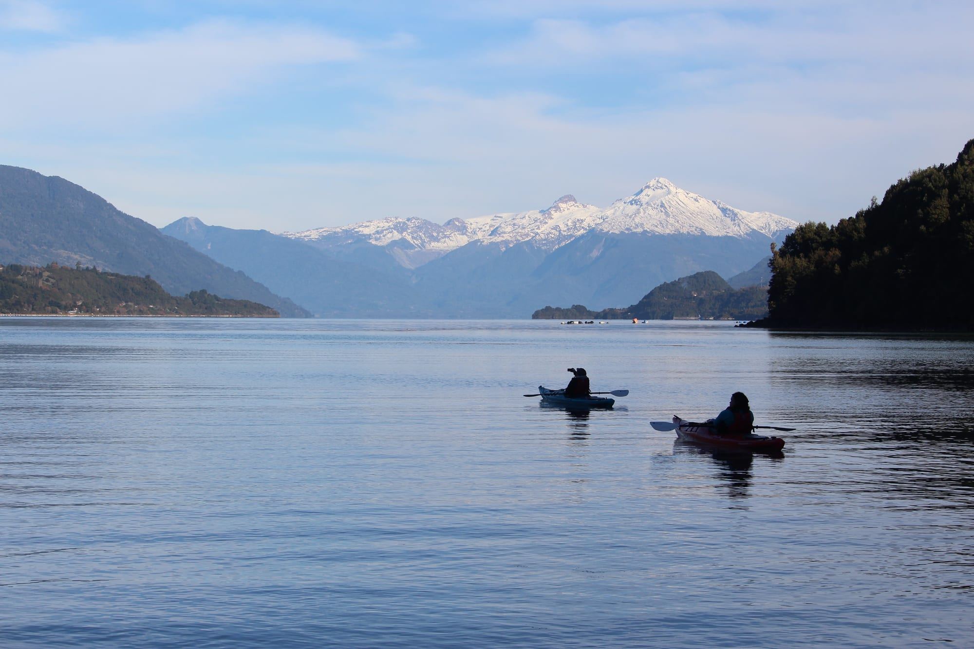 Two kayaks paddle the Reloncaví fjord, with snowy mountains behind