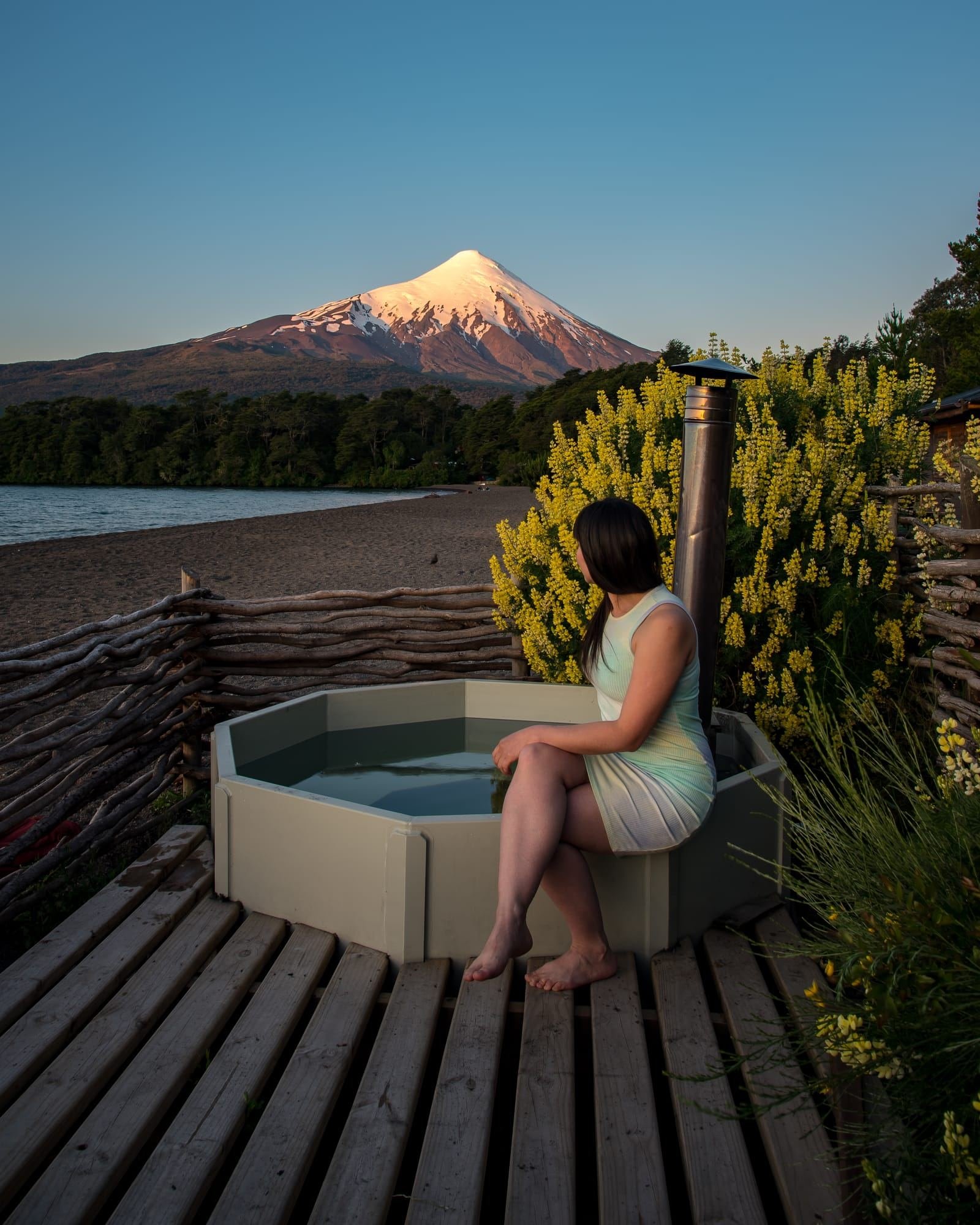 A person sitting on the edge of a hot tub at sunset, with Osorno Volcano behind