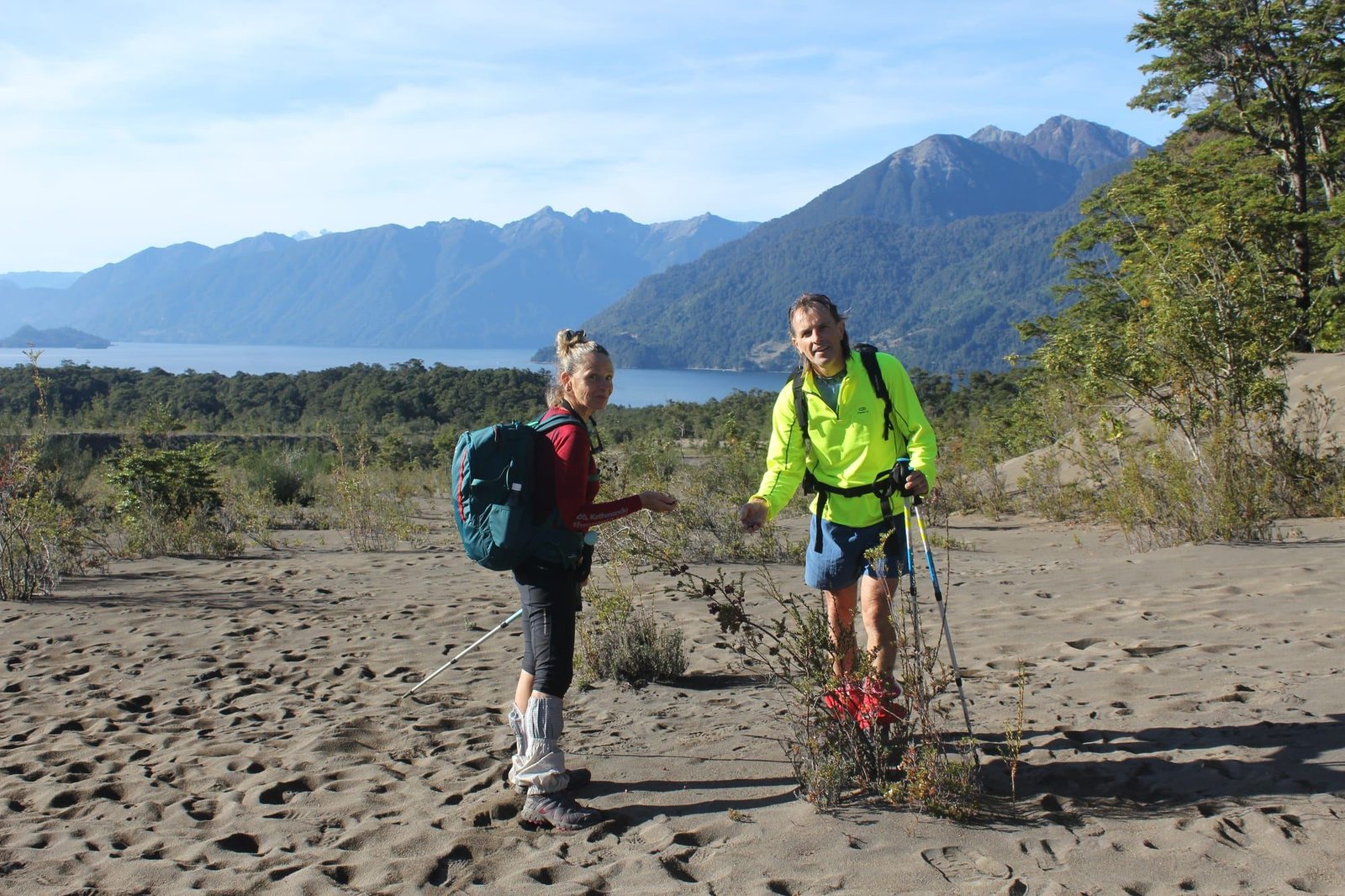 Dos caminantes con bastones en un arenal volcánico, con un lago y montañas al fondo