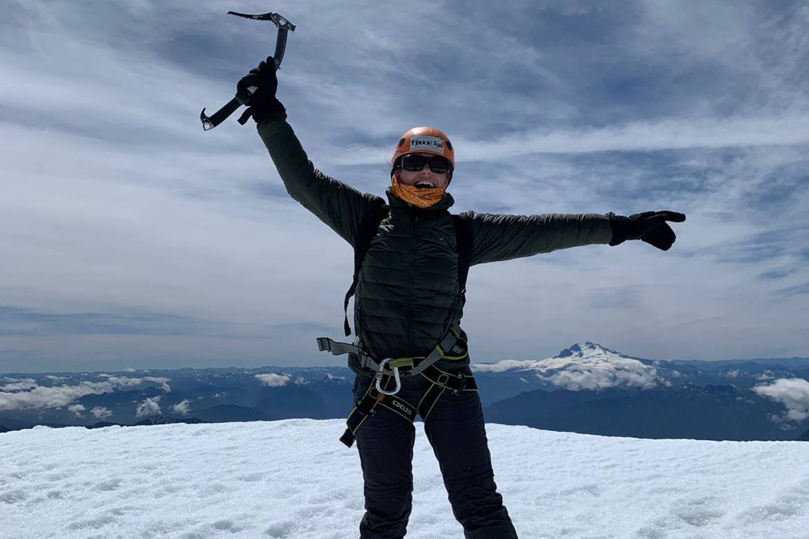 Person in a harness raising an ice axe on the snow, with a snow-capped volcano behind